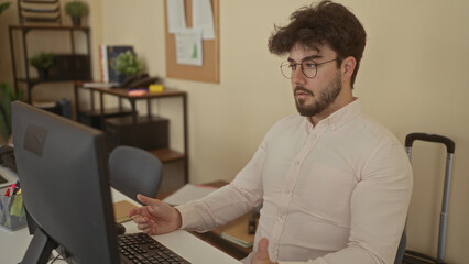 Young man with beard and glasses working on a computer in an office setting, surrounded by shelves and plants, suggesting a focused indoor workspace environment.