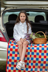 Young girl sitting on a blanket with a basket of flowers at a car trunk during a sunny day in the countryside