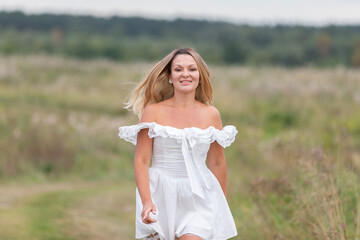 Woman walks joyfully through a grassy field in a white off-shoulder dress on an overcast day