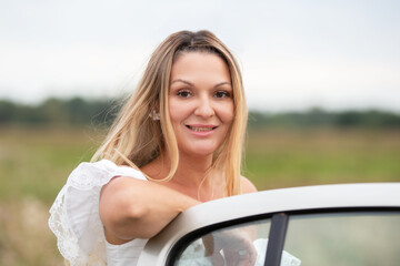 Woman in a white dress leaning on a classic car in an open field during a cloudy day