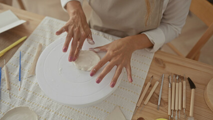 Young blonde woman in apron presses wet clay with bare hands on spinning pottery wheel in artisan...