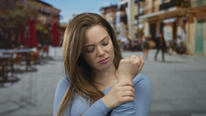 Fototapeta premium Woman in blue sweater frowning holds wrist on busy sunlit street cafe terrace with outdoor tables; discomfort.