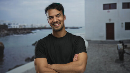 Fototapeta premium Young hispanic man smiling confidently on seaside promenade with arms crossed, serene ocean backdrop enhancing outdoor setting.