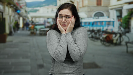 Fototapeta premium Woman with brunette hair shows emotional expressions on an outdoor street with blurred urban scenery, wearing glasses and a grey sweater, exhibiting frustration and contemplation.