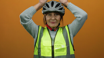 Senior hispanic woman wearing a helmet and safety vest smiling against an orange background, making playful ear gestures above her head, conveying a sense of humor and safety.