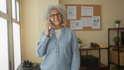 Woman with grey hair stands in office talking on phone, wearing glasses and denim shirt, surrounded by charts and office equipment in a well-lit indoor setting.