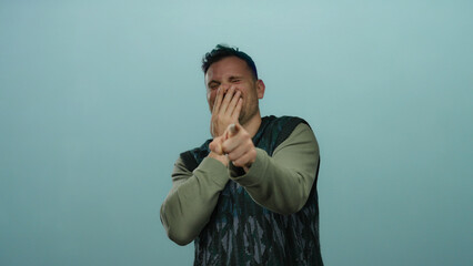 Young man laughing with hand over face and pointing, set against a solid blue background, exuding...