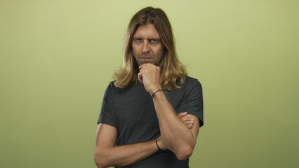 Man with long blond hair and hand on chin slightly leaning in studio against a neutral green wall;...