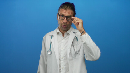 Blond doctor with long hair wearing stethoscope adjusts glasses in studio against blue wall; serious concern.