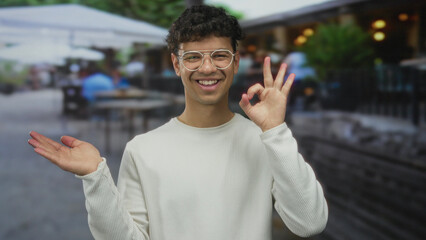 Young man with glasses holds open palm and points finger toward empty space on street cafe terrace;...