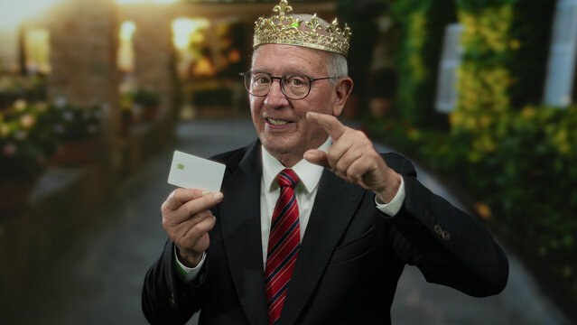 Senior businessman wearing a crown and suit makes a phone gesture while standing on a street, holding a card in an outdoor setting, suggesting a royal announcement. - Powered by Adobe