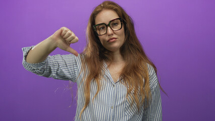 Woman in striped shirt wearing glasses making a thumbs down gesture in a purple studio; disapproval criticism.