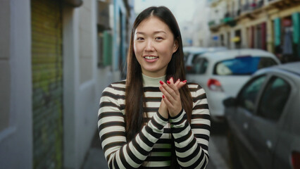 Fototapeta premium Woman clapping on urban street with cars in background, wearing striped sweater, showcasing cheerful expression in city, embodying beauty and youth with charm in an outdoor scene.