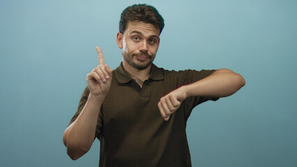 Young hispanic man pointing finger and checking wrist while looking at wrist in light blue studio;...