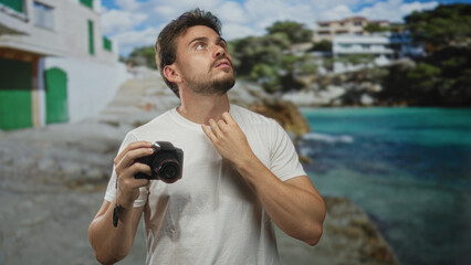 Man holding camera with right hand against seaside backdrop in studio set, looking upward and adjusting collar; contemplation travel memory.