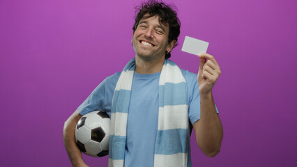 Hispanic man holding a soccer ball and card against a purple background, smiling while wearing a blue shirt and scarf, embodying cheerful sports enthusiasm.