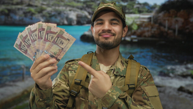 Man in camouflage uniform points finger and smiles at fan of saudi riyal banknotes at street; pride reward.