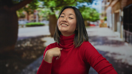 Woman smiling in red sweater points at herself on a vibrant city street, showcasing a self-assured...