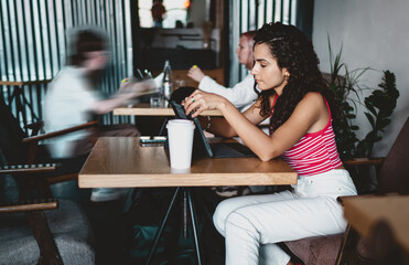 Woman interacts with tablet at café, focused and seated upright, stylus in hand, surrounded by motion and blurred figures, portraying deep concentration amid digital chaos.