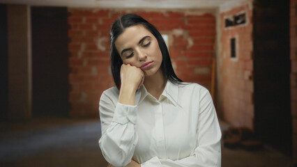 Hispanic woman standing thoughtfully indoors at a construction site, wearing a white shirt with a...