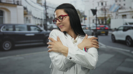 Fototapeta premium Young hispanic woman in scientist uniform embraces herself on an outdoor street, exuding confidence and independence.