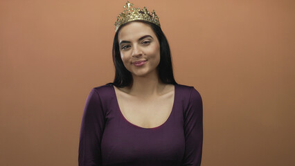 Young hispanic woman with a crown and long black hair smiles warmly against a plain brown wall,...