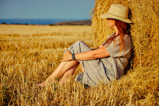 red-haired beautiful woman with hat in field with haystacks against blue sky