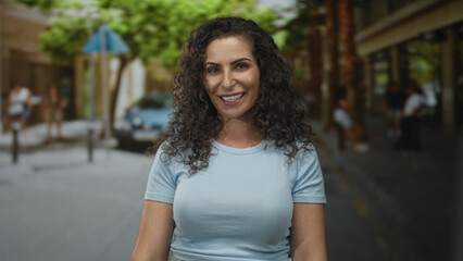 Young hispanic woman face smiling in blue top on sunlit city street with green trees; confidence optimism.