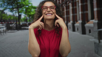 Fototapeta premium Woman wearing red glasses gestures with both index fingers on cheeks in bright sunlight on street; pure happiness.