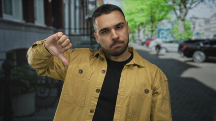 Naklejka premium Man in yellow jacket and black shirt on urban street makes thumbs down gesture with left hand and neutral expression; rejection.