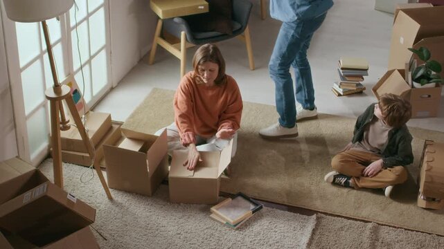 High angle shot of mother and her son sitting on floor and looking at pictures while unpacking, surrounded by cardboard boxes after moving into new home