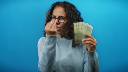 Woman holding argentine pesos angrily against a blue wall isolated as banknotes fan out in her...