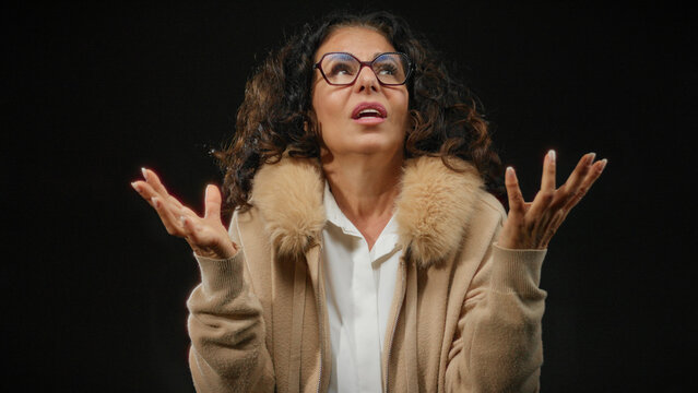 Hispanic woman with curly hair and glasses gesturing expressively against an isolated black background wearing a fur-trimmed coat.