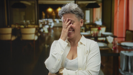 Smiling woman with short hair covers face with hand in building restaurant interior; lighthearted amusement.