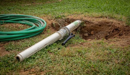 A section of a light gray PVC pipe lies in a dug-out hole in a grassy yard.  A green garden hose is coiled nearby