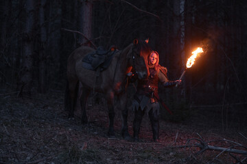 Warrior woman stands with her horse, torch illuminating a dark forest path.