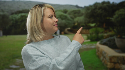 Young blonde woman wearing a light blue sweater points finger to right against green park hedge;...