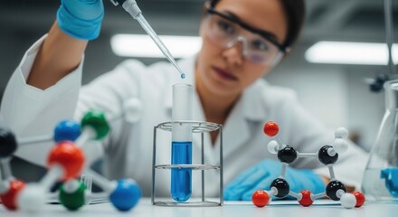 A person in a lab coat and safety goggles uses a pipette to dispense blue liquid into a test tube in a rack with various molecular models and lab glassware on a white surface