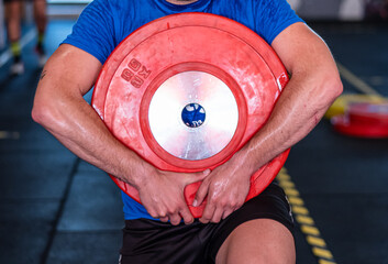 Man in blue shirt and black shorts gripping a heavy red weight plate with both hands, sweating and...