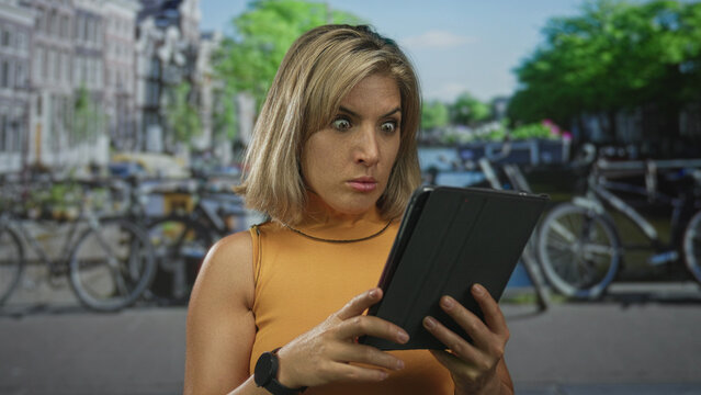 Woman tapping tablet screen with intent focus on an urban bike lined canal street in amsterdam; surprise.