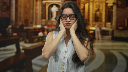 Young hispanic woman pressing ears to head with pained expression in ornate church building...