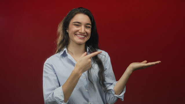 Hispanic teen woman in striped shirt pointing finger to open palm in red studio setting; confidence.