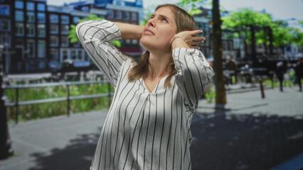 Woman in striped shirt covers ears on busy street beside canal railings under bright sunny sky;...