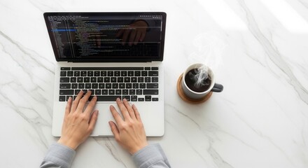 Overhead view of hands typing on a silver laptop displaying code with a steaming dark beverage in a gray mug on a wooden coaster all on a white marble surface