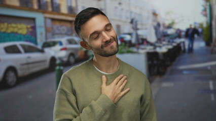 Young hispanic man standing on an urban street with cars and graffiti in the background, placing hand on chest in a friendly gesture.