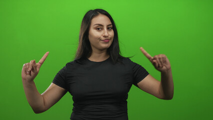Woman in black shirt points both index fingers upward and holds palms open while shrugging in green studio; doubt indecision.