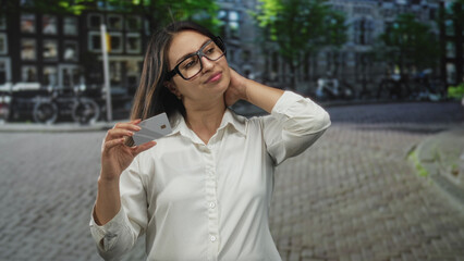 Fototapeta premium Woman holding creditcard close to camera while gesturing with free hand on a cobblestone city street, wearing glasses; payment frustration.