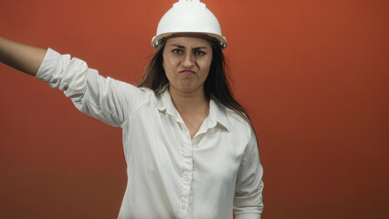 Young woman architect in white hardhat with arms crossed and stern face in orange studio; frustration.