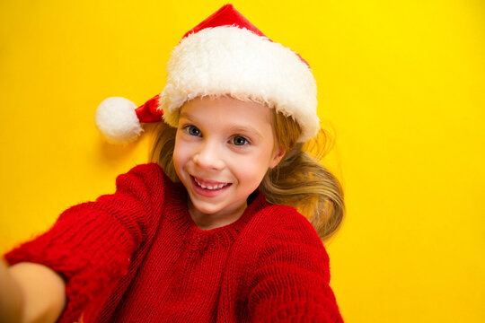 Funny cheerful kid in red sweater wearing Santa hat smiles in bright yellow background for festive holiday selfie