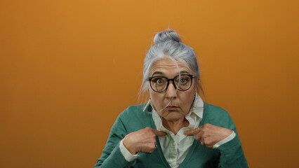 Woman with grey hair and glasses pointing at herself, wearing a green cardigan against an orange...